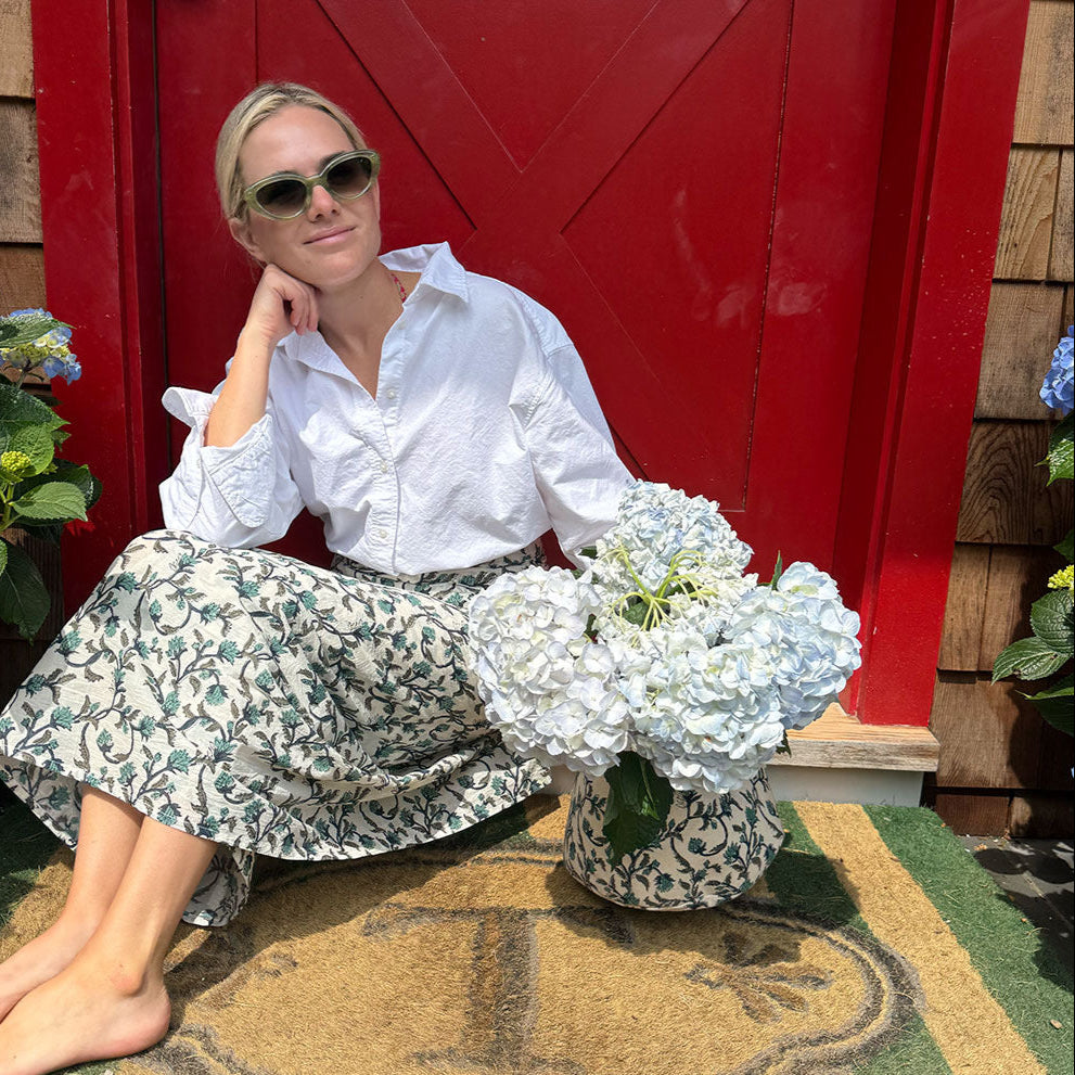 Woman in white shirt and floral skirt sitting on a mat with a red door and plants in the background