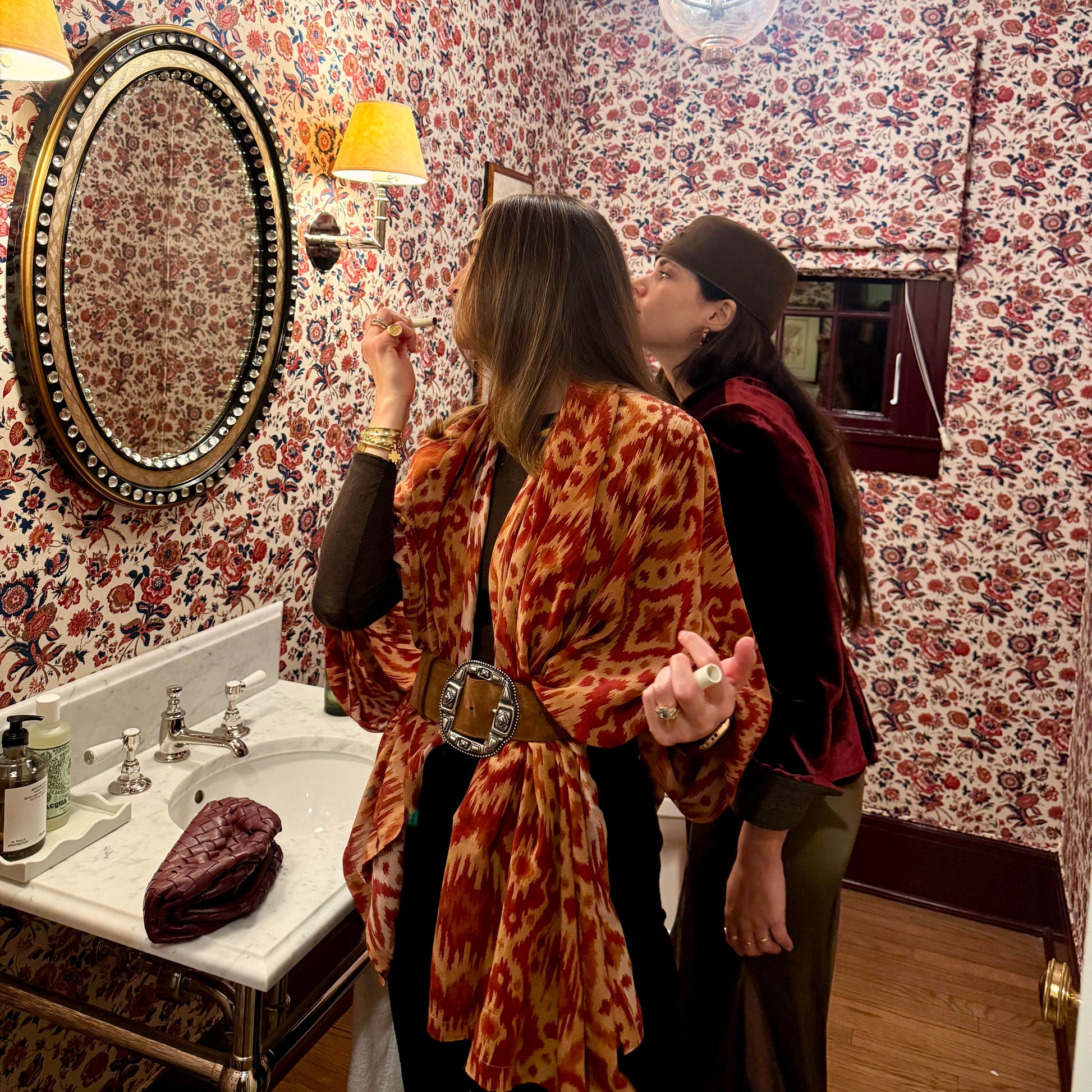Two women in a bathroom with floral wallpaper and a round mirror.